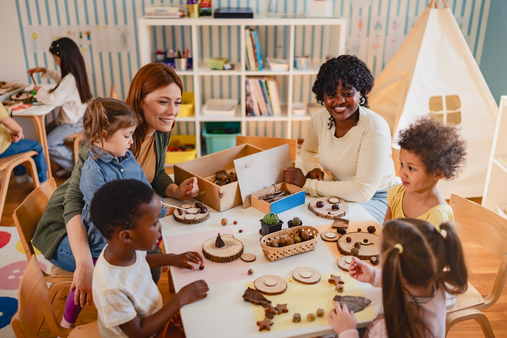 Diverse group of preschool children engage in a nature-based learning activity with two teachers, using wood and natural objects to explore creativity and tactile skills in the classroom.